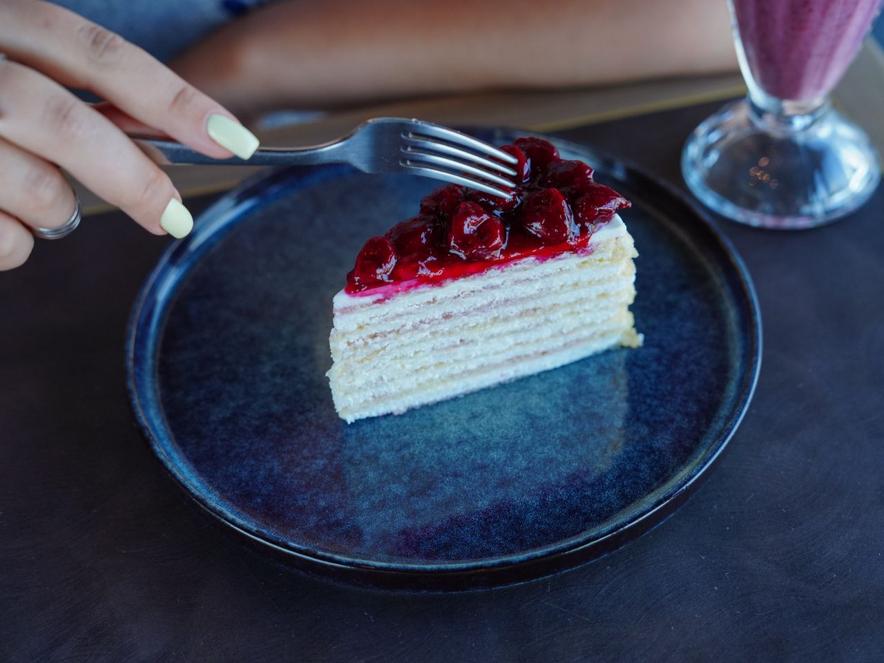 A fork held by a woman captures a cherry-topped cake slice on a blue plate, showcasing dessert indulgence.